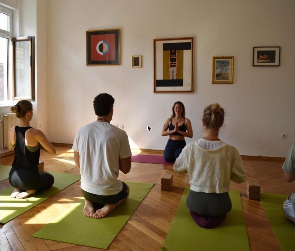 Skupinska praksa joge v studiu, učiteljica sedi pred skupino z dlanmi v namaste